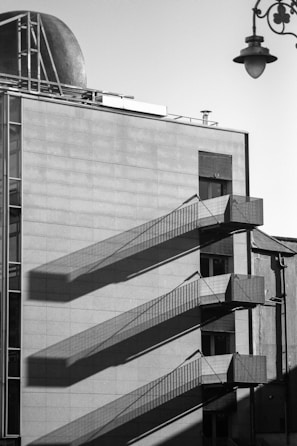 A monochromatic, industrial-style building facade with external metal staircases casting long, geometric shadows on the flat, tiled surface. A metallic dome and a vintage streetlamp add contrast to the modern architectural lines.