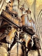 An intricately designed pipe organ with elaborate wooden carvings, situated in what appears to be a historical cathedral or church. The architecture features high vaulted ceilings and stone walls, with decorative statues and detailed woodwork surrounding the organ pipes.