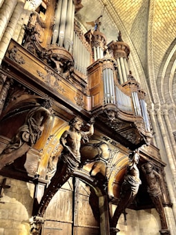 An intricately designed pipe organ with elaborate wooden carvings, situated in what appears to be a historical cathedral or church. The architecture features high vaulted ceilings and stone walls, with decorative statues and detailed woodwork surrounding the organ pipes.