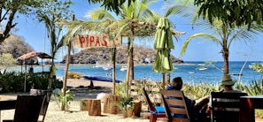 A tropical beach scene with lush palm trees and a wooden sign reading 'PIPAS' hanging overhead. The view extends to a sandy beach where kayaks and boats are seen, along with distant rocky cliffs. Two people are relaxing in lounge chairs under the shade, enjoying the coastal atmosphere.