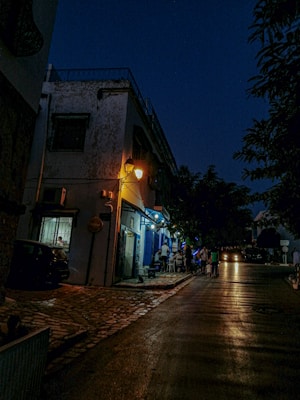 A narrow cobblestone street is dimly lit by a warm streetlamp, creating a cozy atmosphere. A small group of people is gathered outside a blue-themed caf&eacute;, which has an inviting glow from its interior lights. The building exterior shows signs of wear, adding a rustic charm. The sky is dark, suggesting nighttime, and trees line the sidewalk, casting shadows on the ground.