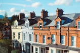 A row of traditional terraced houses with red brick facades, featuring multiple chimneys atop the steep, slate-tiled roofs. The houses have white-framed windows and a decorative facade, typical of urban residential architecture. A satellite dish is attached to one of the buildings.