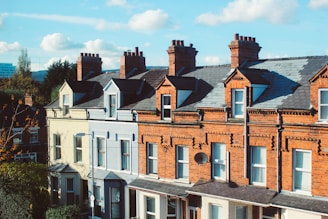 A row of traditional terraced houses with red brick facades, featuring multiple chimneys atop the steep, slate-tiled roofs. The houses have white-framed windows and a decorative facade, typical of urban residential architecture. A satellite dish is attached to one of the buildings.