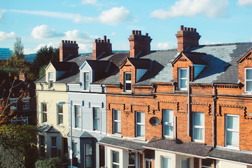 A row of traditional terraced houses with red brick facades, featuring multiple chimneys atop the steep, slate-tiled roofs. The houses have white-framed windows and a decorative facade, typical of urban residential architecture. A satellite dish is attached to one of the buildings.