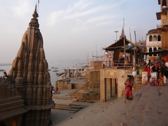 A historical riverside scene featuring a large, intricately detailed temple structure with a dome-like top made of stone. The background shows a river with several small boats and a haze typical of early morning or late afternoon. There are people in traditional attire walking along steps that lead towards the water, suggesting a cultural or religious gathering. The architecture is characteristic of an ancient or heritage site.