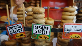 Stacks of bagels are displayed on rods behind three price signs. The signs label the bagels with humorous names such as 'Enough Already,' 'Sesame Street,' and '1610 Traditional,' and each is priced at $1.50. A person is partially visible in the background.