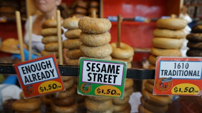 Stacks of bagels are displayed on rods behind three price signs. The signs label the bagels with humorous names such as 'Enough Already,' 'Sesame Street,' and '1610 Traditional,' and each is priced at $1.50. A person is partially visible in the background.