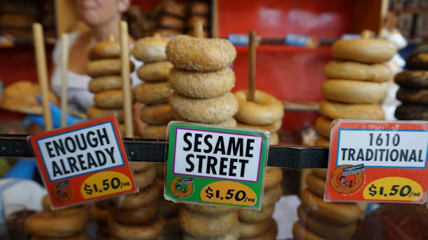 Stacks of bagels are displayed on rods behind three price signs. The signs label the bagels with humorous names such as 'Enough Already,' 'Sesame Street,' and '1610 Traditional,' and each is priced at $1.50. A person is partially visible in the background.