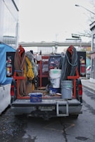 A utility truck is parked on the street, its bed filled with various equipment and tools. Coiled cables are neatly arranged on either side, with a range of colors including red, yellow, and black. Buckets and containers are also present, suggesting preparation for a construction or maintenance task. A large welding machine is visible in the middle, accompanied by fire extinguishers mounted on the truck.