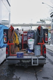 A utility truck is parked on the street, its bed filled with various equipment and tools. Coiled cables are neatly arranged on either side, with a range of colors including red, yellow, and black. Buckets and containers are also present, suggesting preparation for a construction or maintenance task. A large welding machine is visible in the middle, accompanied by fire extinguishers mounted on the truck.