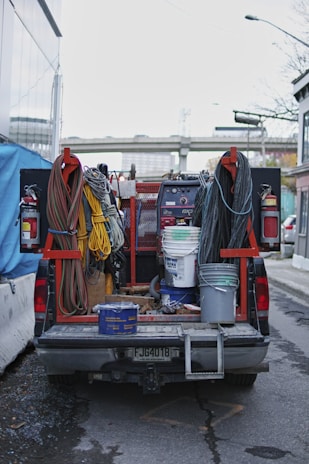 A utility truck is parked on the street, its bed filled with various equipment and tools. Coiled cables are neatly arranged on either side, with a range of colors including red, yellow, and black. Buckets and containers are also present, suggesting preparation for a construction or maintenance task. A large welding machine is visible in the middle, accompanied by fire extinguishers mounted on the truck.