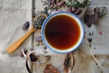 A ceramic cup filled with dark tea is placed on a textured fabric background. Surrounding the cup are an assortment of dried tea leaves, seeds, and spices, including pieces of bark and cacao beans. A wooden spoon holds some of the tea leaves on the side.
