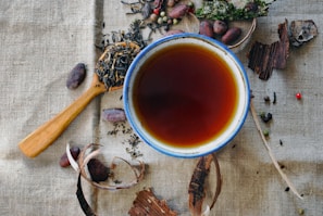 A ceramic cup filled with dark tea is placed on a textured fabric background. Surrounding the cup are an assortment of dried tea leaves, seeds, and spices, including pieces of bark and cacao beans. A wooden spoon holds some of the tea leaves on the side.
