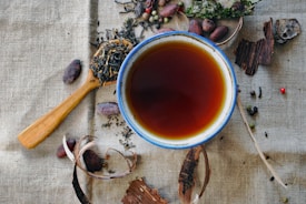 A ceramic cup filled with dark tea is placed on a textured fabric background. Surrounding the cup are an assortment of dried tea leaves, seeds, and spices, including pieces of bark and cacao beans. A wooden spoon holds some of the tea leaves on the side.
