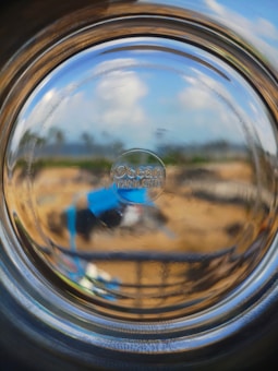 A view through the bottom of a glass bottle, distorting the scenery behind it. The background includes a blurred outdoor setting with earthy tones and some blue elements. The text 'Ocean Thailand' is embossed on the glass, focused in the center of the image.