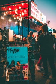 A vibrant street food stall is illuminated by a string of hanging lights, creating a warm and inviting atmosphere. A glowing sign displays text in a foreign language, possibly indicating the type of cuisine offered. Several people, casually dressed, are gathered around the stall, some interacting with the vendor. In the foreground, a reflective surface with the words 'your intuition brought you here' adds a contemporary touch, and a skateboard leans against it, suggesting a youthful vibe.