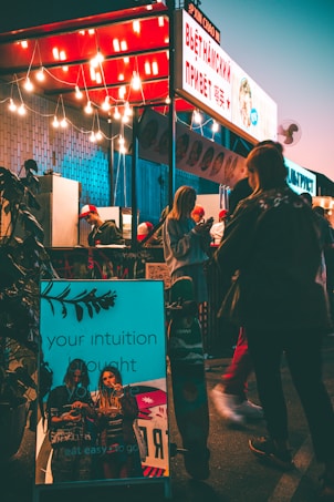 A vibrant street food stall is illuminated by a string of hanging lights, creating a warm and inviting atmosphere. A glowing sign displays text in a foreign language, possibly indicating the type of cuisine offered. Several people, casually dressed, are gathered around the stall, some interacting with the vendor. In the foreground, a reflective surface with the words 'your intuition brought you here' adds a contemporary touch, and a skateboard leans against it, suggesting a youthful vibe.