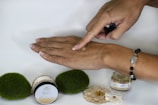 A hand is applying a substance, likely a body balm, on the back of the other hand. There are two small jars near the hand, labeled "Ginger Honey Hemp Body Balm." The background is simple and there are decorative elements like green moss-like spheres and small flowers on the surface. The person is wearing a bracelet with colorful beads.