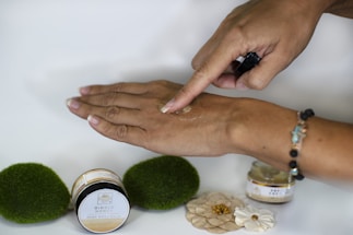 A hand is applying a substance, likely a body balm, on the back of the other hand. There are two small jars near the hand, labeled "Ginger Honey Hemp Body Balm." The background is simple and there are decorative elements like green moss-like spheres and small flowers on the surface. The person is wearing a bracelet with colorful beads.