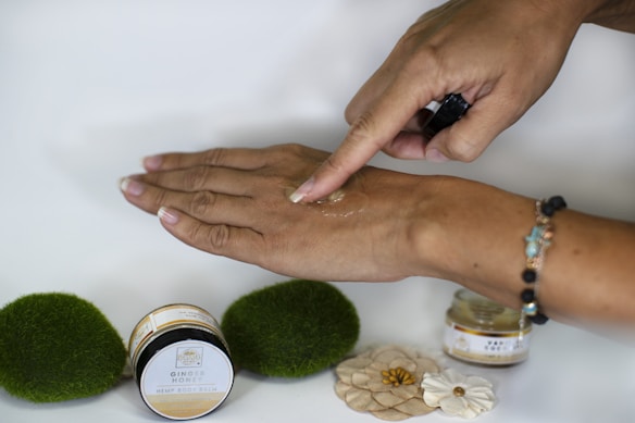 A hand is applying a substance, likely a body balm, on the back of the other hand. There are two small jars near the hand, labeled "Ginger Honey Hemp Body Balm." The background is simple and there are decorative elements like green moss-like spheres and small flowers on the surface. The person is wearing a bracelet with colorful beads.