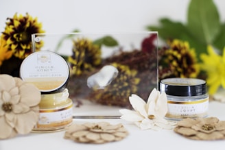 Two jars of hemp body balm are displayed, labeled with ginger honey and vanilla coconut varieties. They are surrounded by decorative dried flowers and leaves with hues of yellow, brown, and green. The setup appears to be on a light surface, creating a natural and earthy aesthetic.