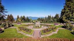 A beautifully manicured garden with vibrant green lawns, colorful flower beds, and symmetrically placed hedges. A central pathway and ornate fountain add elegance to the landscape, with tall trees and mountains visible in the background under a clear blue sky.