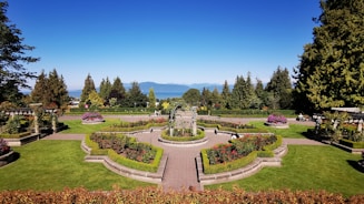 A beautifully manicured garden with vibrant green lawns, colorful flower beds, and symmetrically placed hedges. A central pathway and ornate fountain add elegance to the landscape, with tall trees and mountains visible in the background under a clear blue sky.