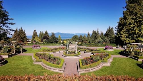 A beautifully manicured garden with vibrant green lawns, colorful flower beds, and symmetrically placed hedges. A central pathway and ornate fountain add elegance to the landscape, with tall trees and mountains visible in the background under a clear blue sky.