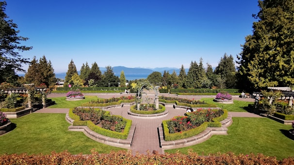 A beautifully manicured garden with vibrant green lawns, colorful flower beds, and symmetrically placed hedges. A central pathway and ornate fountain add elegance to the landscape, with tall trees and mountains visible in the background under a clear blue sky.