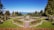 A beautifully manicured garden with vibrant green lawns, colorful flower beds, and symmetrically placed hedges. A central pathway and ornate fountain add elegance to the landscape, with tall trees and mountains visible in the background under a clear blue sky.