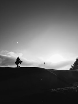 A silhouette of a person carrying golf clubs walks on a hilly golf course. The sun is low, casting a dramatic light and elongating shadows. A flag is visible on the right against a backdrop of soft clouds and a clear sky.