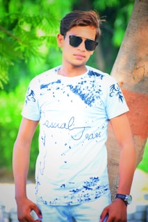 A person wearing a white t-shirt with blue graphic designs and text, paired with light blue jeans. The individual is leaning against a tree, wearing sunglasses. The background is filled with lush green leaves, creating a vibrant and natural setting.