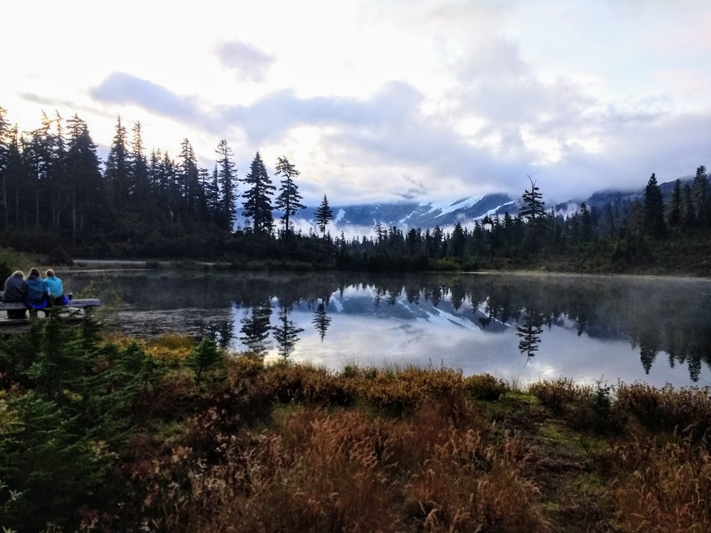 three person sitting on bench beside lake