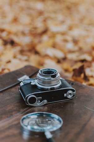 A vintage camera rests on a wooden surface with a blurred, autumnal background of fallen leaves. A magnifying glass is placed nearby, adding to the nostalgic and investigative feel.