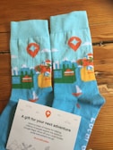 A pair of blue socks with colorful patterns including mountains, trees, a sailboat, and a red map marker. Beside the socks is a card with text expressing gratitude and encouragement, adorned with small illustrated icons.