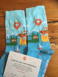 A pair of blue socks with colorful patterns including mountains, trees, a sailboat, and a red map marker. Beside the socks is a card with text expressing gratitude and encouragement, adorned with small illustrated icons.