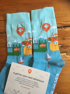 A pair of blue socks with colorful patterns including mountains, trees, a sailboat, and a red map marker. Beside the socks is a card with text expressing gratitude and encouragement, adorned with small illustrated icons.