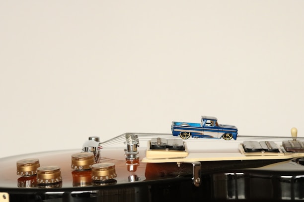 A miniature blue toy truck is positioned on the strings of an acoustic guitar, creating a juxtaposition of scale between the toy and the instrument. The guitar body is a rich brown color with the golden tuning pegs and some parts of the bridge visible.