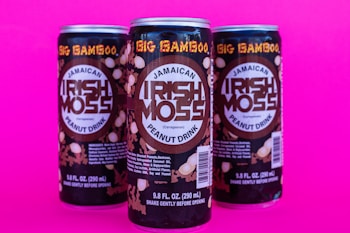 Three cans of Jamaican peanut drink labeled 'Irish Moss' are displayed against a vibrant pink background. The cans have a dark design with the product name in large, bold letters and additional descriptive text below.