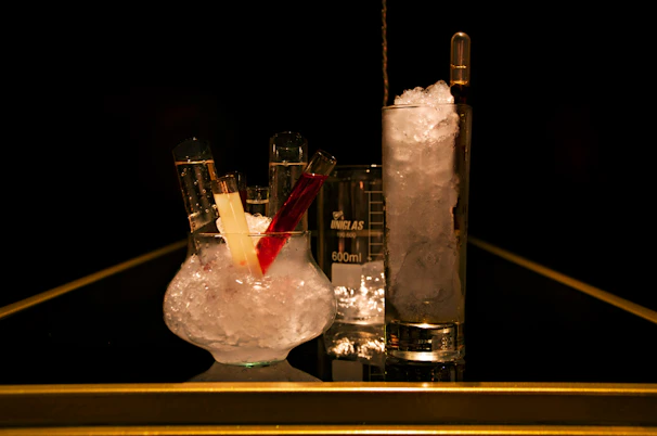 A dark setting with a clear beaker and a cylindrical glass filled with ice, accompanied by a smaller glass container holding crushed ice and three test tubes with colorful liquids, all placed on a reflective surface.