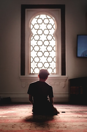 A person is kneeling on the floor inside a quiet, dimly lit room with an ornate window that allows soft, natural light to filter through. The patterned window frame casts intricate shadows on the surrounding surfaces. The atmosphere is serene and contemplative.