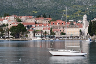 A picturesque coastal town with stone buildings and red-tiled roofs sits at the edge of a calm body of water. A white sailboat rests on the tranquil water, and lush greenery covers the hillside in the background. A clock tower stands out among the buildings, contributing to the historic charm of the scene.