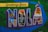 A vibrant mural featuring the phrase 'Greetings from NOLA' against a colorful background. The letters 'NOLA' are filled with various iconic images of New Orleans, including a mardi gras mask, a church, skyline silhouettes, the fleur-de-lis symbol, and a stadium. A pelican is perched on top of the letter 'A'.