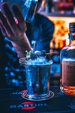 A bartender is pouring a liquid ingredient into a metallic cocktail cup filled with crushed ice and garnished with berries and mint. The scene is set in a dimly lit bar, with a bottle partially visible next to the cup. The lighting creates a vibrant, colorful atmosphere with blue and orange hues.