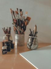 A collection of art supplies is arranged on a wooden table. Various paintbrushes are stored in tin and glass containers. Several small glass bottles of ink or paint are nearby, and an empty canvas lies on the table.