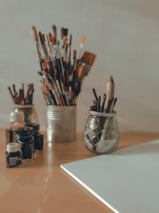A collection of art supplies is arranged on a wooden table. Various paintbrushes are stored in tin and glass containers. Several small glass bottles of ink or paint are nearby, and an empty canvas lies on the table.