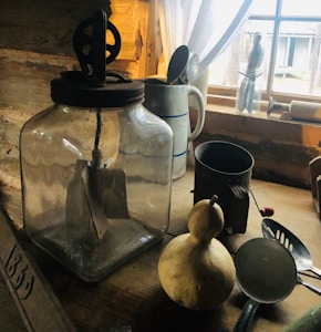 The image features an assortment of rustic kitchen items arranged on a wooden surface. A large glass jar with a metal gear mechanism sits prominently, accompanied by an aged metal pitcher with a handle, a vintage iron scoop, and a gourd. A wooden window frame allows soft natural light to filter in, illuminating the textured wood grains and highlighting the rustic charm of the objects.