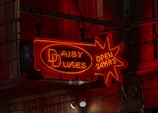 A neon sign displaying the words 'Daisy Dukes' and 'Open 24 Hrs' in a starburst pattern, with additional text offering breakfast, lunch, and dinner. The sign emits a warm red and orange glow against a dark, industrial background with faintly visible windows.