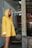 A person wearing a bright yellow raincoat with a hood stands near a rustic wooden structure. The setting appears to be outdoors, with elements of weathered wood and a partially visible poster in the background. The person looks contemplative, gazing off to the side.