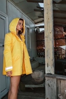 A person wearing a bright yellow raincoat with a hood stands near a rustic wooden structure. The setting appears to be outdoors, with elements of weathered wood and a partially visible poster in the background. The person looks contemplative, gazing off to the side.